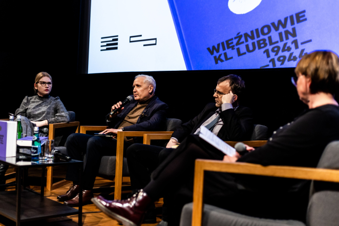 Cztery siedzące osoby na zdjęciu. Jedna z nich trzyma mikrofon. W tle plansza z okładką książki