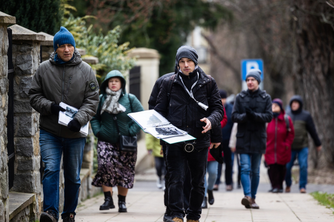 Grupa osób idzie przez miasto. W środku mężczyzna, który trzyma w dłoni segregator ze zdjęciami