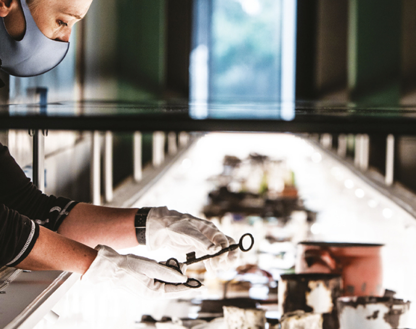 Kobieta w maseczce  i białych bawełnianych rękawiczkach trzyma w dłoniach stary klucz, dookoła przedmioty