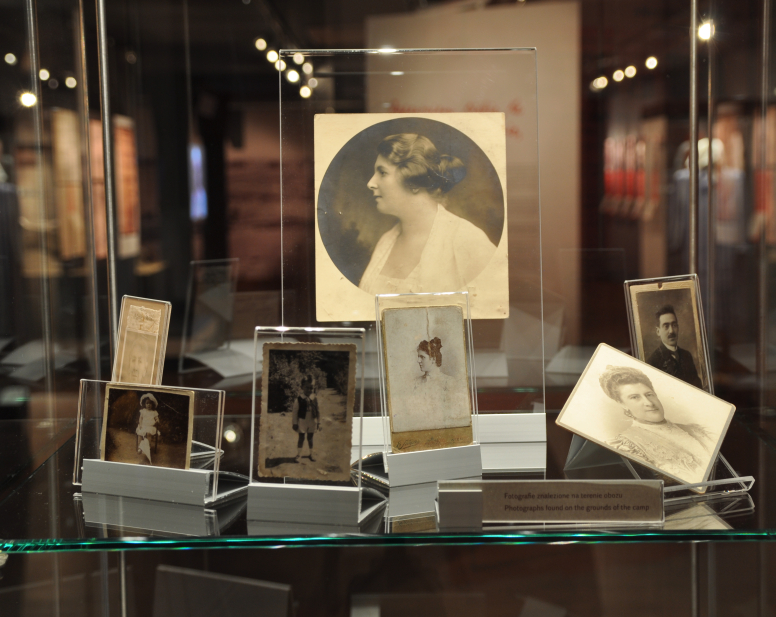 Zdjęcie szklanej gabloty, w której stoją czarno-białe zdjęcia prezentujące twarze lub sylwetki ludzi