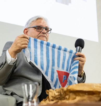 Mężczyzna o siwych włosach trzyma chustę w biało-niebieskie pasy czerwony trójkąt z literą P. 