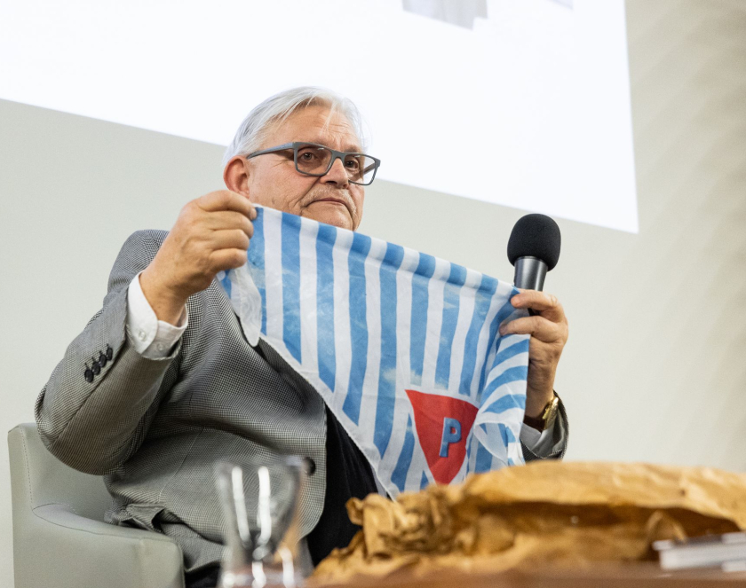 Mężczyzna o siwych włosach trzyma chustę w biało-niebieskie pasy czerwony trójkąt z literą P.