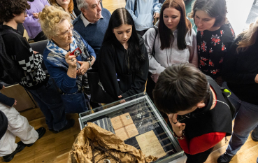 Grupa osób nad przeszkloną gablotą, młodzież oraz starsza kobieta w biało-niebieskiej chuście. 