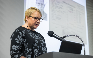 Kobieta o jasnych, krótkich włosach przemawia do mikrofonu, na drugim planie widoczna część prezentacji.