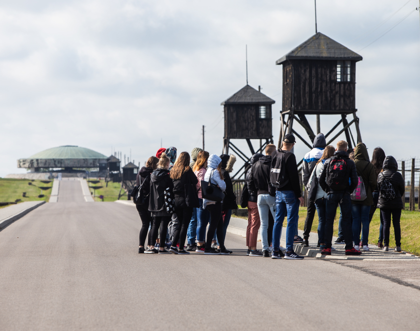 Grupa młodych ludzi stoi na skraju jezdni. Za nimi wieżyczka wartownicza, a dalej pomnik z betonową kopułą