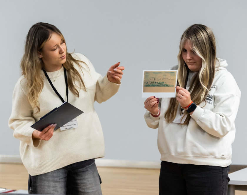 Dwie kobiety stoją w sali i omawiają reprodukcję historycznego rysunku. Jedna z nich wskazuje na obraz, trzymając w ręku notatnik, a druga prezentuje kartę z ilustracją przedstawiającą krajobraz lub zabudowania.