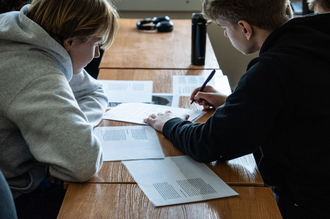 Dwóch nastoletnich mężczyzn przy szkolnych ławkach uzupełnia materiały na zadrukowanych kartkach A4. 