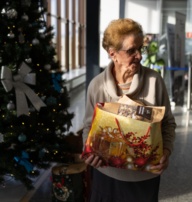 Starsza kobieta stojąca pod choinką trzyma w rękach paczkę 