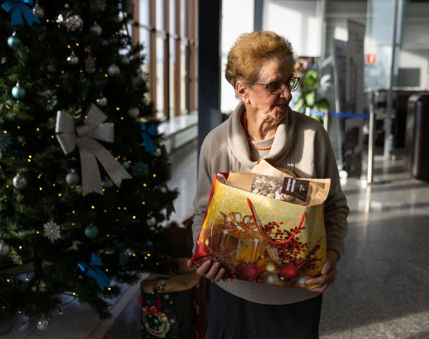Starsza kobieta stojąca pod choinką trzyma w rękach paczkę 
