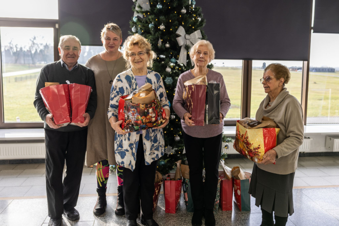 Grupa osób: trzy starsze kobiety, jedna w średnim wieku i jeden starszy mężczyzna, stoją obok choinki trzymają w rękach paczki 