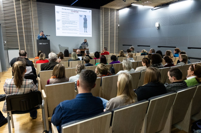 Grupa osób w sali audytoryjnej, na scenie prelegentka Urszula Cur. 