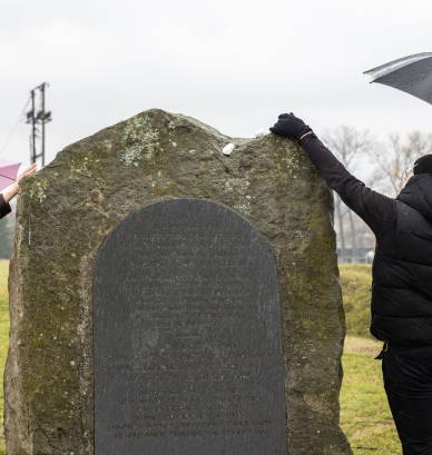 Kobieta i mężczyzna w zimowych ubraniach kładą kamienia na kamień-pomnik, oboje trzymają parasole nad głowami