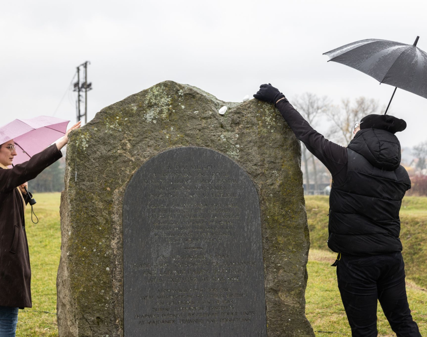 Kobieta i mężczyzna w zimowych ubraniach kładą kamienia na kamień-pomnik, oboje trzymają parasole nad głowami