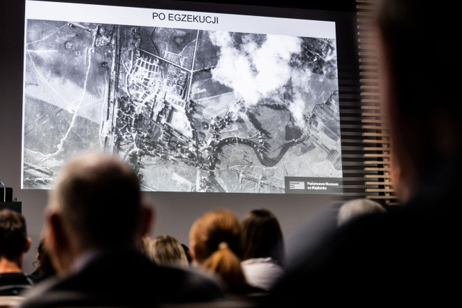 Grupa osób na sali audytoryjnej, na ekranie kinowym prezentacja: zdjęcie lotnicze i napis: po egzekucji 