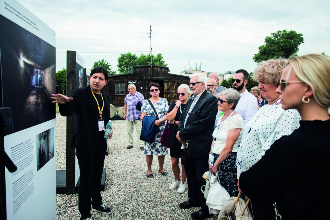 Po lewej tablice z wystawą i opowiadający o nich mężczyzna. Po prawej grupa ludzi w różnym wieku