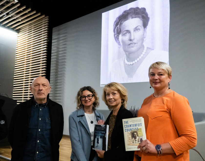 Trzy kobiety oraz łysy mężczyzna na tle prezentacji z czarno-białym zdjęciem kobiety o upiętych włosach. 