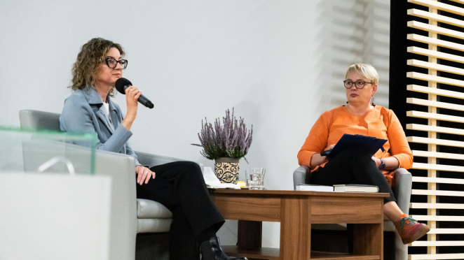 Dwie kobiety na scenie, kobieta z kręconymi włosami przemawia do mikrofonu. 