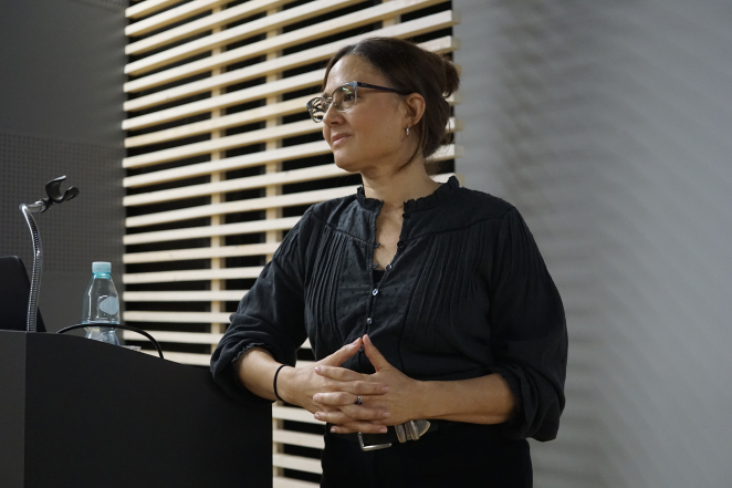 Kobieta z ciemnymi włosami i okularami stoi przy czarnym pulpicie.