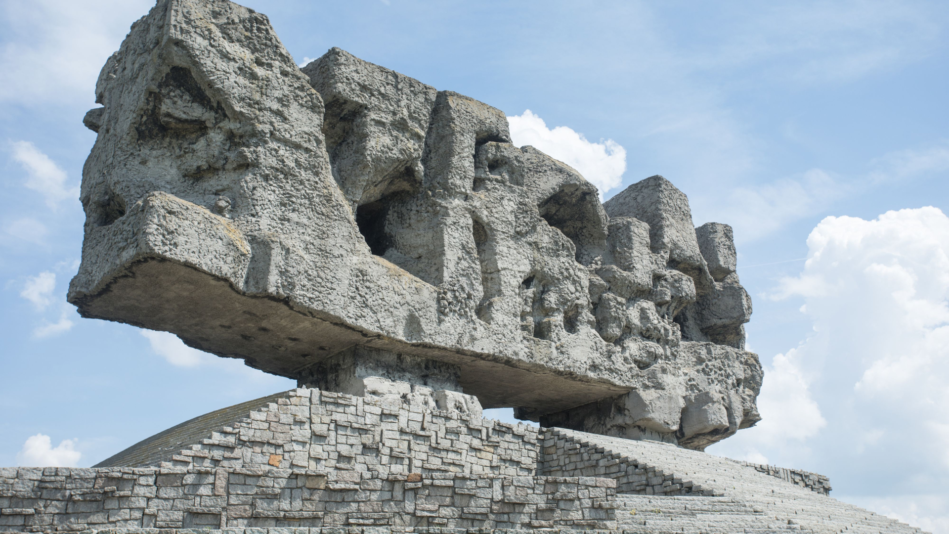 Zdjęcie pomnika-bramy na Majdanku. Bryła pomnika w formie prostokąta z kamieni nachodzących na siebie. Widok z dołu. W tle niebo. 