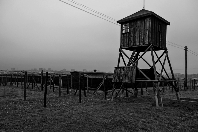 Czarno-białe zdjęcie. Drewniana wieżyczka wartownicza, za nią ogrodzenie z drutu i baraki