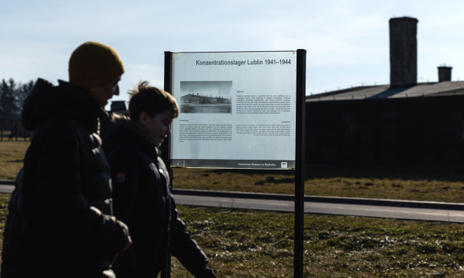 Szklana tablica ze ścieżki historycznej, na pierwszym planie dwójka zwiedzających, na drugim barak więźniarski. 