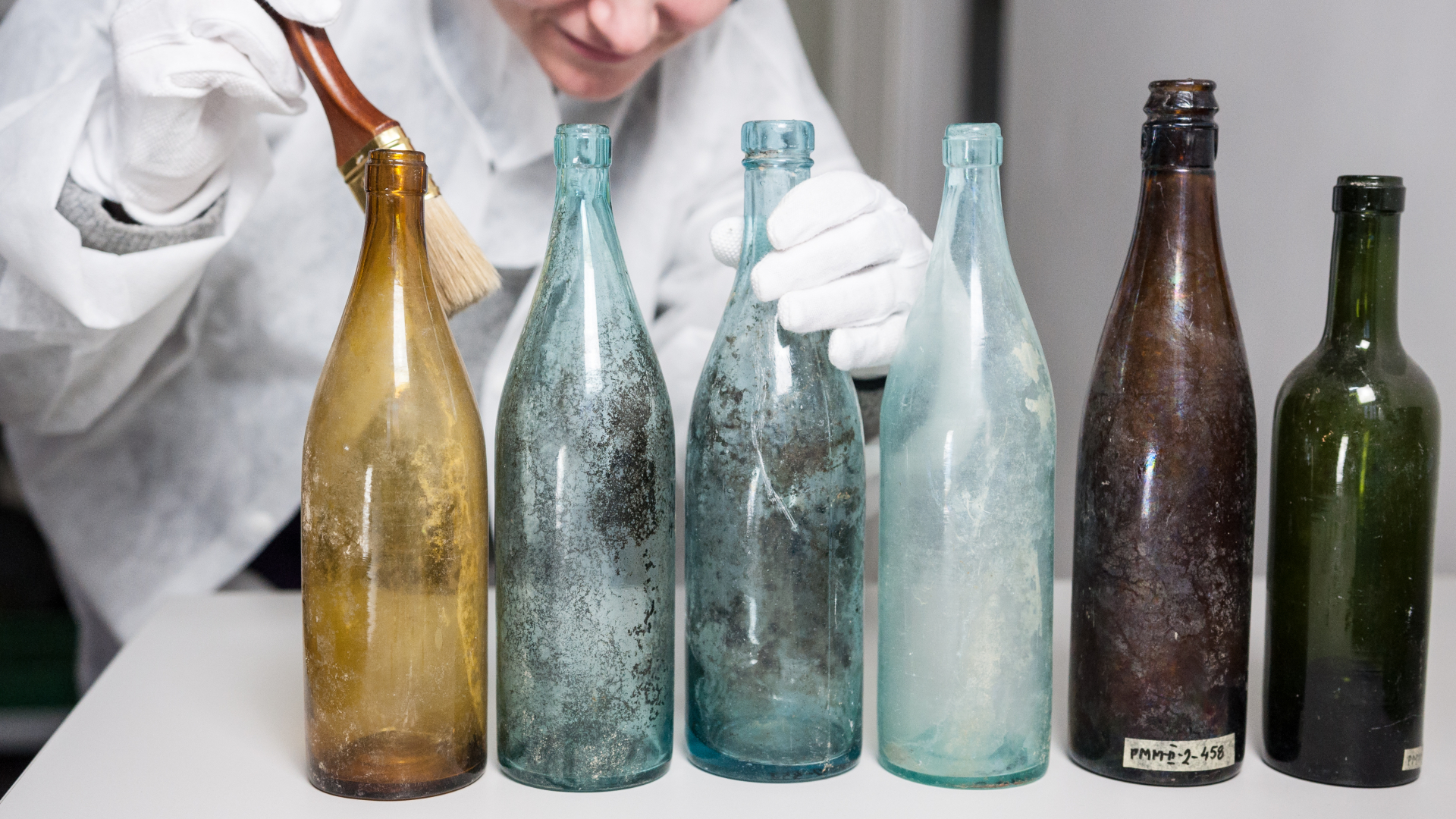 Kobieta w białym stroju i białych rękawiczkach oczyszcza pędzelkiem sześć kolorowych, szklanych butelek.