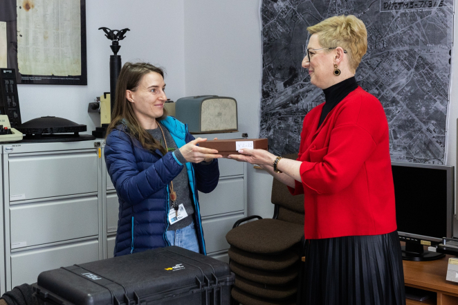 Dwie kobiety przekazujące sobie brązowe pudełko z białą naklejką, jedna z kobiet ubrana w czerwony żakiet, druga w lekką puchową granatową kurtkę, na drugim planie pracowania archiwum: mapa, przedmioty zabytkowe oraz repliki pomników wykonane techniką druku 3d