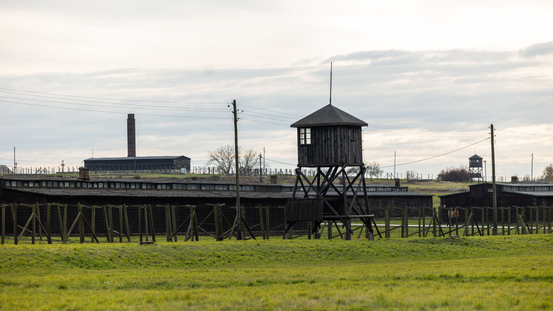 Zdjęcie współczesne z widokiem na obóz. Od lewej baraki, krematorium i wieża strażnicza, barak. W tle w oddali kolejna wieża. 