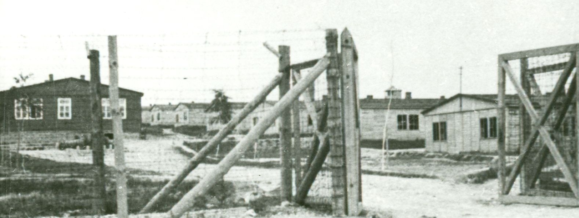 Brama z drutu kolczastego oraz drewna, w tle rzędy baraków więźniarskich 