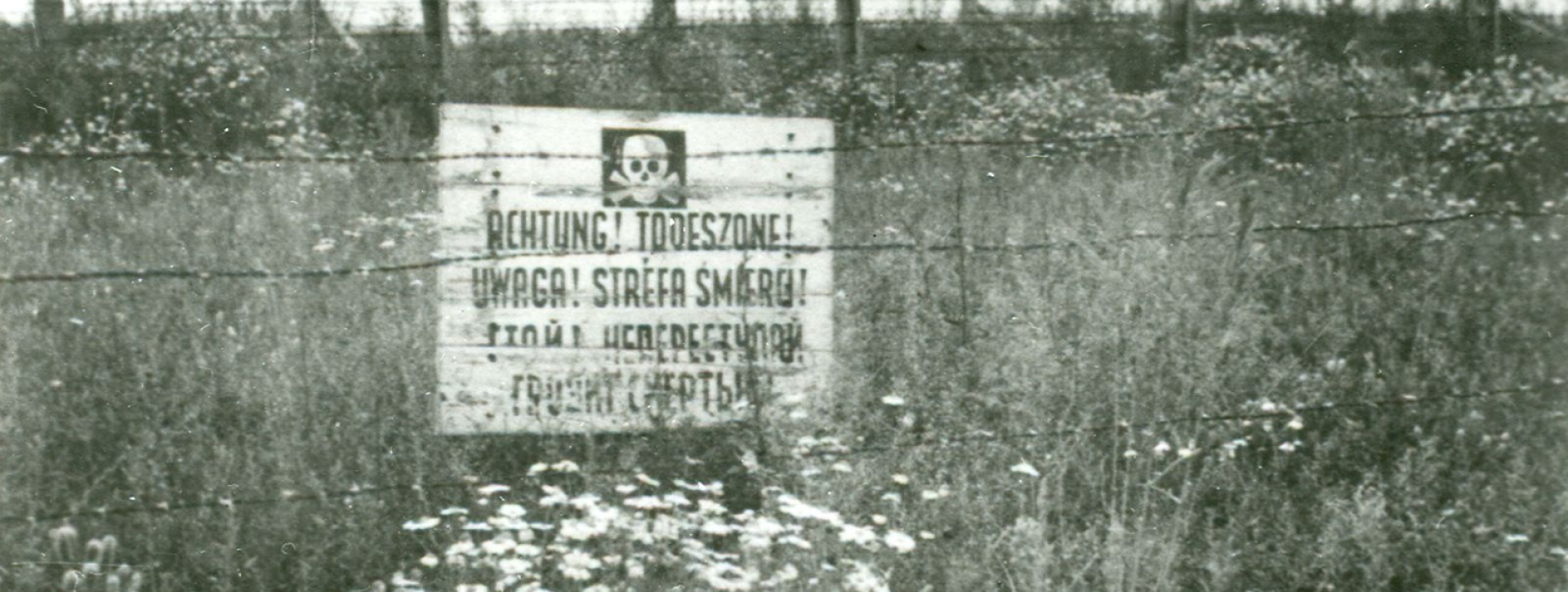Ogrodzenie z drutów kolczastych oraz drewnianych belek, w ziemie wbita biała tablica z napisem: Achtung! Todeszone!Uwaga! Strefa Śmierci! dalsza treść słabo widoczna 