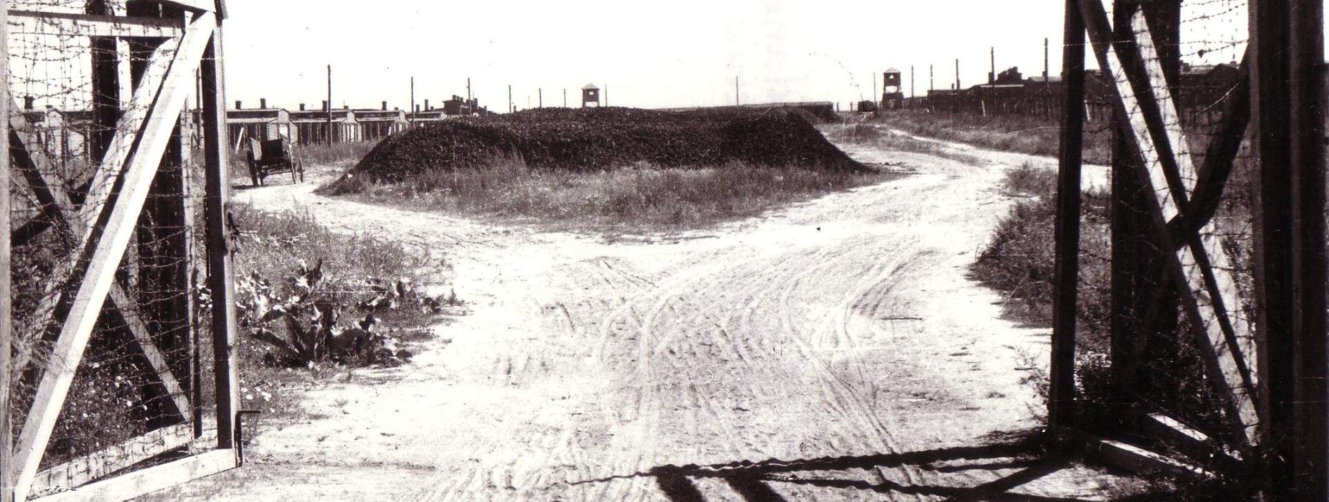 Otwarta brama, pośrodku usypany kopiec ziemi, w tle zabudowa obozowa 