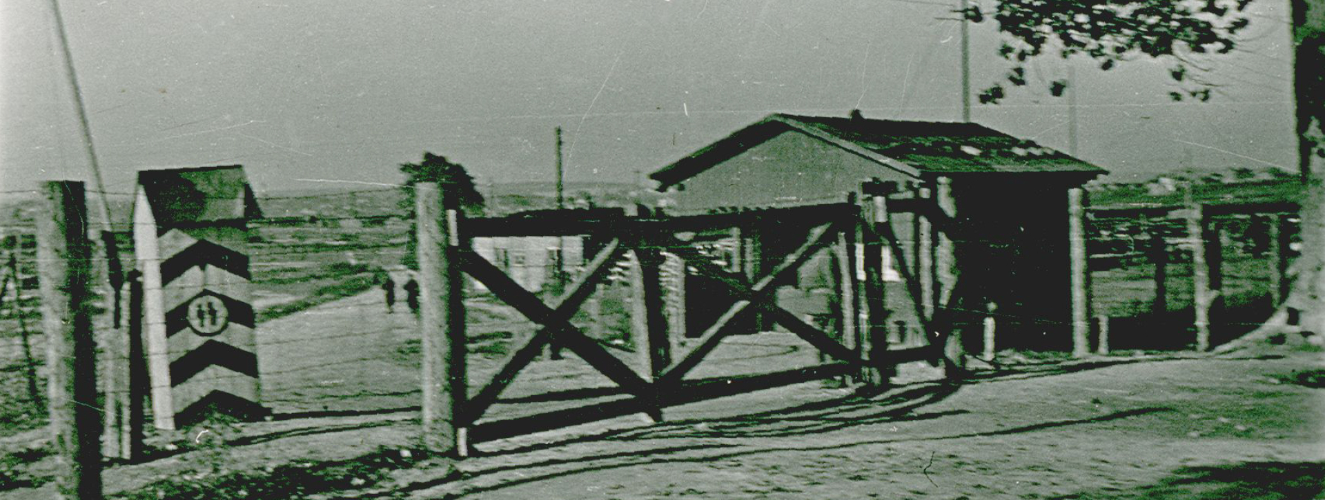 Brama wjazdowa, po lewej budka strażnicza z symbolem nazistowskim, po prawej niska drewniana zabudowa wartownicza 