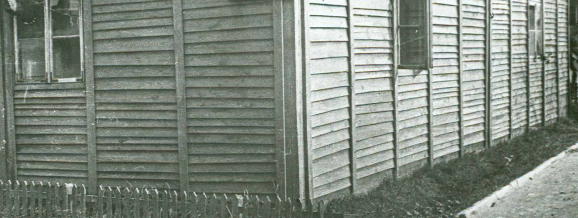 Drewniany barak z trzema oknami, na dłuższym boku widoczna tabliczka BLOCK 2 