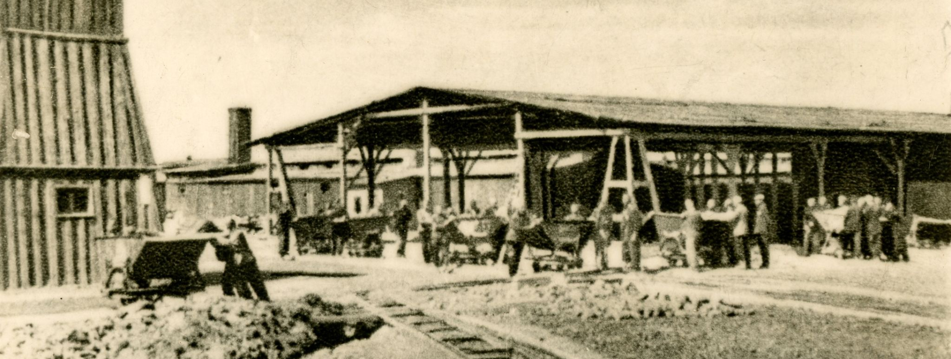 Grupy mężczyzn podczas pracy, część osób przy kilku wagonikach kolejowych, w tle dużych rozmiarów drewniana wiata, po lewo fragment wieży wartowniczej 