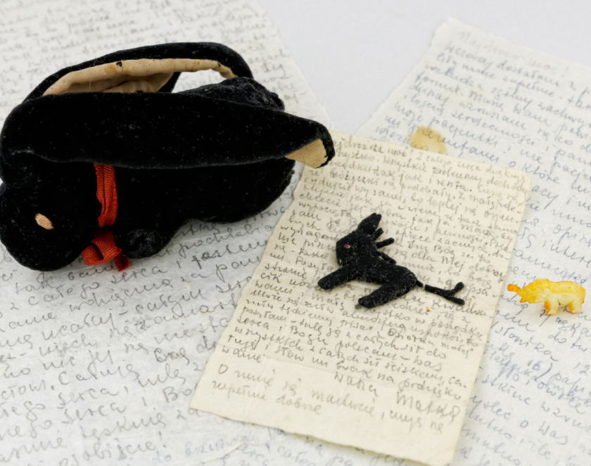 Pluszowa czarna maskotka z długimi uszami, obok mała przypominająca konia oraz słoń z kości słoniowej, dookoła odręcznie napisane listy