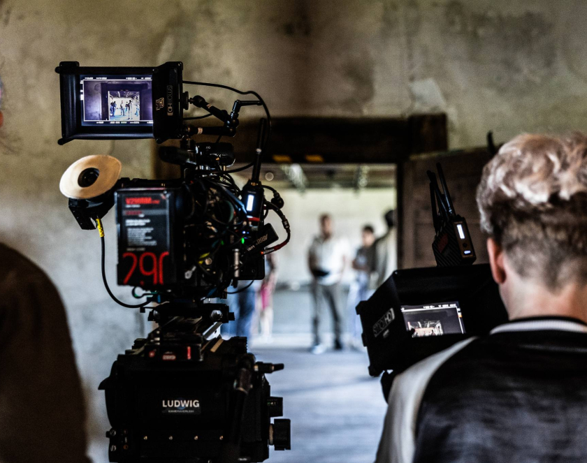 Dwie osoby stoją we wnętrzu budynku odwrócone plecami. Przed nimi kamery. W tle niewyraźne postaci.