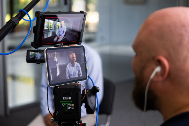 Na zdjęciu wyświetlacze kamery i telefonu, na których widać mężczyznę w jasnym ubraniu. Po prawej głowa łysego mężczyzny ze słuchawką w uchu