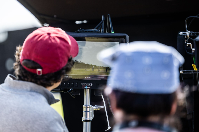 Zdjęcie. Dwie osoby stoją odwrócone plecami. Przed nimi wyświetlacz kamery. Na nim niewyraźne postaci obok baraków więźniarskich.