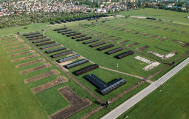 Zdjęcie z drona, teren obozu koncentracyjnego na Majdanku, kilka pół więźniarskich, ślady po barakach, jedno z pół z obozową zabudową, na drugim planie zabudowa miejska 