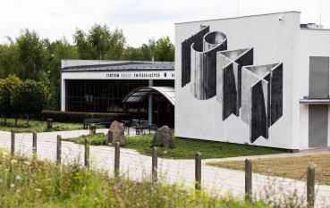 Budynek Centrum obsługi Zwiedzających,  na pierwszym planie ogrodzenie z drutów kolczastych, na drugim budynek z muralem: 1944, wejście z półokrągłym daszkiem i napisem Centrum Obsługi Zwiedzających, przed budynkiem dwa kamienie-pomniki 