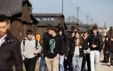 Grupa młodzieży, po lewej stronie rzędy baraków więźniarskich 