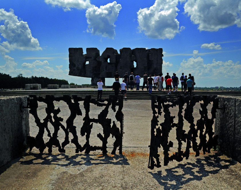 Grupa zwiedzających przechodzi przez metalową bramę z sylwetkami postaci i kieruje się w stronę monumentalnego Pomnika Walki i Męczeństwa na terenie byłego obozu koncentracyjnego na Majdanku, pod błękitnym niebem z chmurami.
