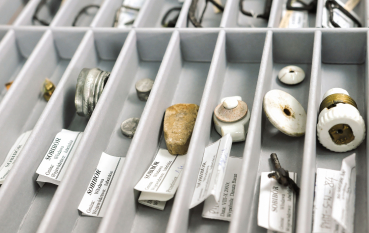 Zbiór drobnych przedmiotów w pudełku z przegródkami, widoczne numery inwentarzowe i napis: Sobibór 