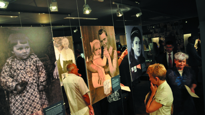 wiedzający oglądają ekspozycję muzealną z dużymi, wiszącymi fotografiami portretowymi przedstawiającymi osoby z przeszłości; zdjęcia są podświetlone i rozmieszczone w ciemnej sali wystawowej