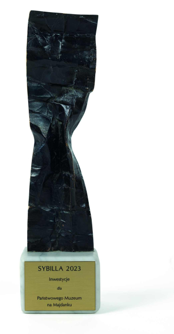 Statuetka nagrody Sybilla 2023 o abstrakcyjnej, ciemnej formie, umieszczona na jasnej podstawie z tabliczką z napisem „Sybilla 2023 – Inwestycje dla Państwowego Muzeum na Majdanku”