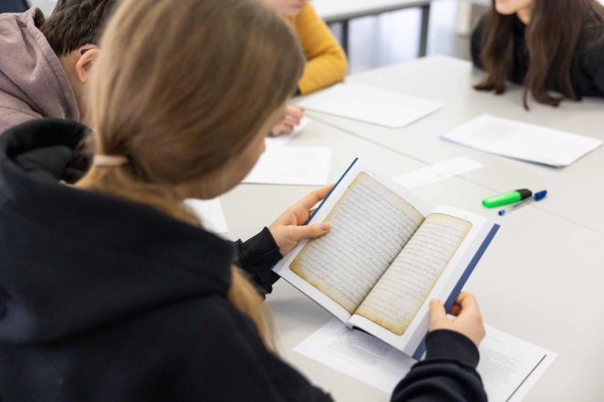 Uczestniczka zajęć siedzi przy stole i czyta otwartą książkę podczas warsztatów edukacyjnych; w tle widoczne są inne osoby oraz materiały do pracy.