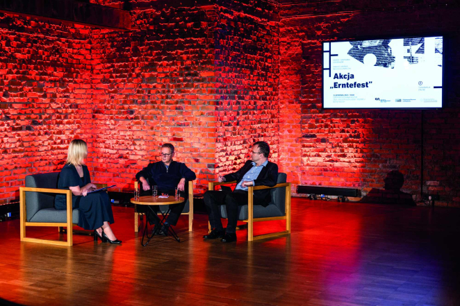 Panel dyskusyjny odbywający się w sali z ceglaną ścianą podświetloną na czerwono; trzy osoby siedzą w fotelach i prowadzą rozmowę na scenie.