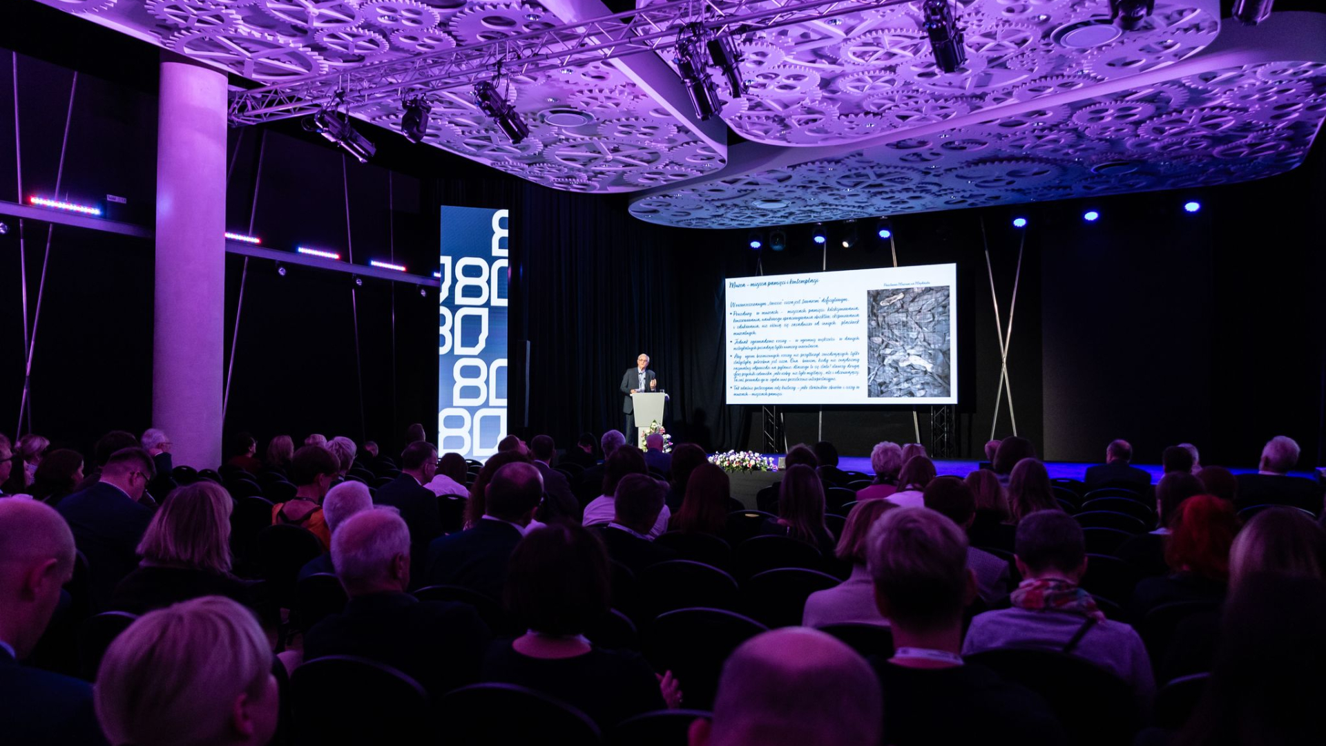 Prelegent przemawia z mównicy podczas konferencji w sali audytoryjnej; publiczność siedzi na widowni, a za mówcą widoczny jest ekran z prezentacją