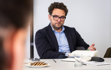 Mężczyzna w średnim wieku w okularach o kręconych włosach ubrany w garnitur, na pierwszym planie otwarty notes.