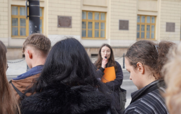 Grupa młodych osób w tle młoda kobieta w okularach przemawia do mikrofonu.